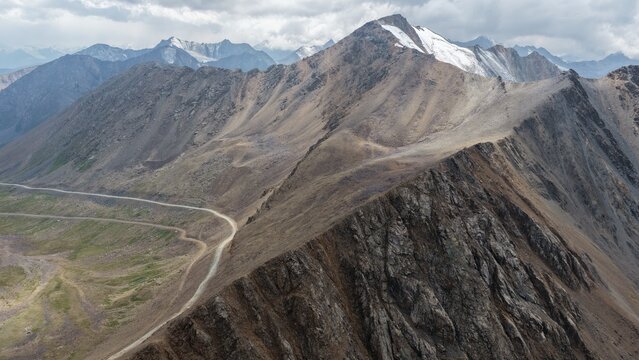 Aerial view of the rugged Enilchek mountain range in the Tian Shan, featuring a winding dirt road cutting through rocky terrain under a cloudy sky in Enilchek, Issyk-Kul Region, Kyrgyzstan.