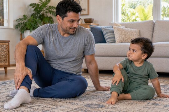 Father Sitting On The Living Room Floor With Grumpy Toddler Son