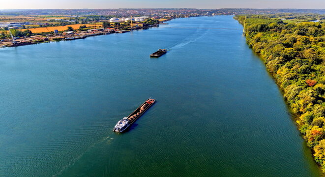 Aerial view of the Danube river with cargo barges navigating past the Smederevo shipyard and industrial facilities along the lush green riverbanks in Smederevo, Podunavlje District, Serbia.