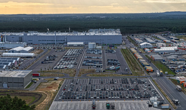 Aerial view of Tesla Gigafactory Berlin-Brandenburg, showing the manufacturing facility, parking lots filled with new cars, and surrounding forest in Grunheide Mark, Brandenburg, Germany.