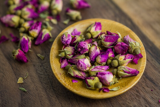 Close-up of dried rose buds, some on a small wooden plate, others scattered on a dark wooden surface, used for tea or decoration.