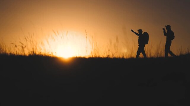 Business team silhouette hill. Two hikers walk along the hill at sunset, with the sun setting behind them. The hillside features tall grass as the two hikers explore the peaceful landscape.