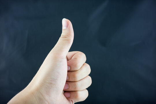 A close-up of a human hand making a thumbs up gesture, set against a dark plain background, conveying approval and positivity.