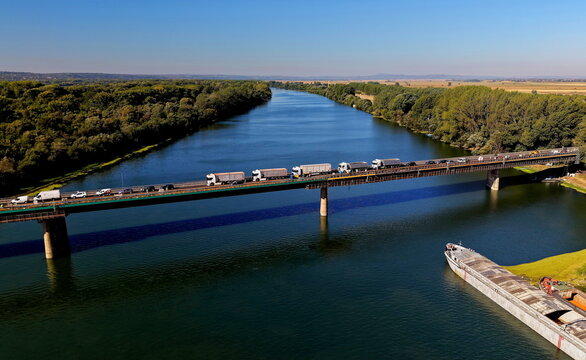 Aerial view of the bridge over the Danube River with trucks crossing and a barge moored below, surrounded by lush riverbanks under a clear blue sky in Smederevo, Podunavlje District, Serbia.