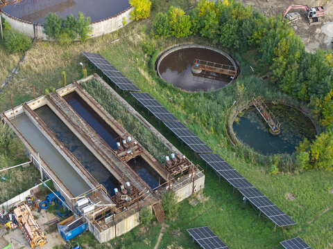 Aerial view of a wastewater treatment plant featuring circular sedimentation tanks, rectangular aeration basins, and rows of solar panels set within a green landscape in Poland.