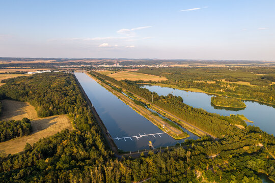 Aerial view of Labe Arena Racice surrounded by lush green forests and calm water under a clear summer sky in Racice, Ustecky kraj, Czechia.