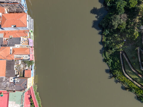 Aerial view of a river flowing past a row of colorful buildings with terracotta roofs on one bank and a lush green park with winding pathways on the other in Brazil.