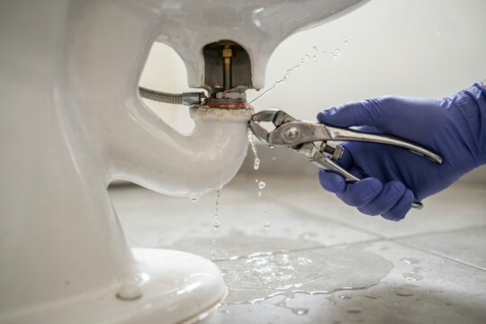 Professional plumber in blue gloves using pliers to repair leaking toilet bowl base with water spraying on bathroom floor.