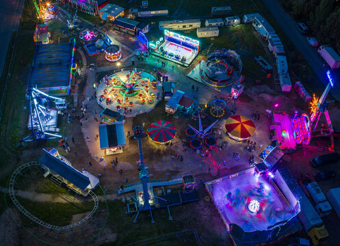 Aerial view of a vibrant night fairground featuring illuminated amusement rides, colorful tents, and parked trailers under the night sky in Vilnius, Vilnius County, Lithuania.