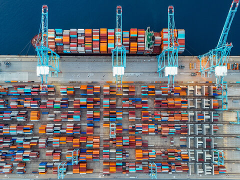 Aerial view of the semi-automated container port with large cranes, stacks of colorful shipping containers, and a cargo ship docked at the pier in Vado Ligure, Liguria, Italy.