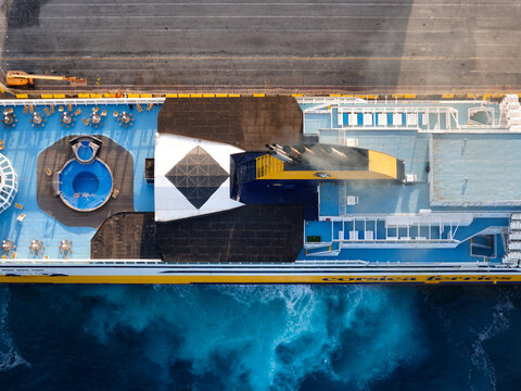 Aerial view of a Corsica Ferries ship deck with a swimming pool, lounge chairs, and smoke rising from the funnel, sailing on the blue sea in Vado Ligure, Liguria, Italy.
