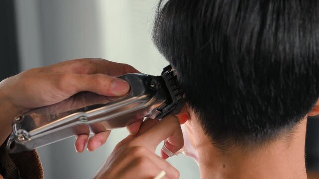 A barber uses electric clippers to trim a seated customer&rsquo;s haircut. The barber guides the metal clipper teeth along dark hair while the customer wears a face mask and protective cape.