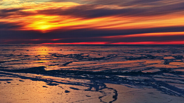 Aerial view of the frozen sea surface with cracked ice patterns reflecting the vibrant orange and red colors of a sunset in Latvia.