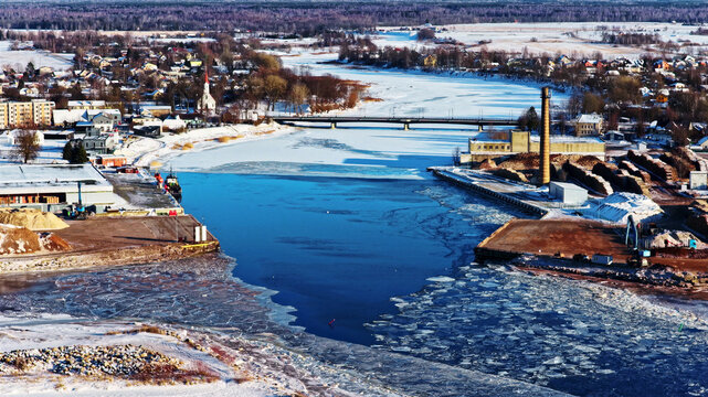 Aerial view of a partially frozen river with ice floes flowing past an industrial site featuring a tall brick chimney, with a bridge and a small town in the background in Latvia.