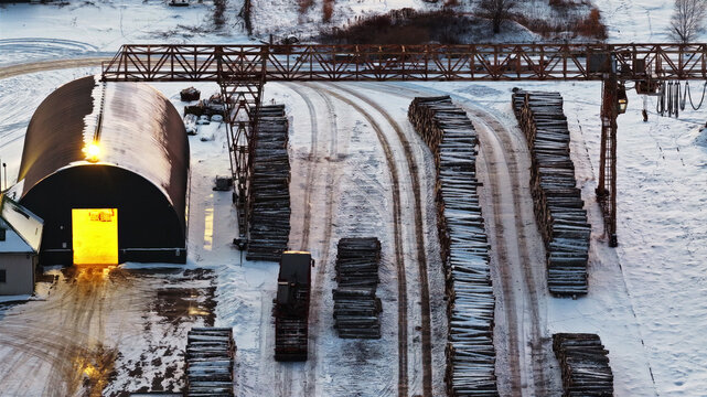 Aerial view of a snow-covered lumber yard featuring stacks of logs, a large gantry crane, and a dark arched storage building with a glowing entrance in Latvia.