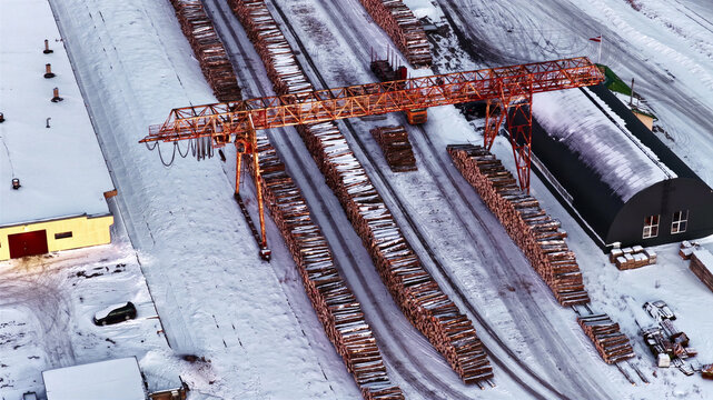 Aerial view of a lumber yard with large stacks of logs and a gantry crane in a snowy winter landscape in Latvia.