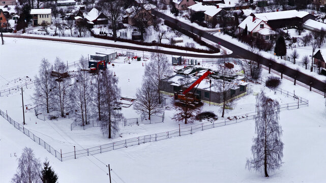 Aerial view of a construction site featuring a crane and building structure surrounded by snow-covered ground and bare trees in a rural landscape in Latvija.