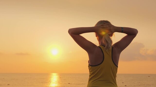 Woman stretches arms and breathes deeply while looking at sunrise over tropical sea.
