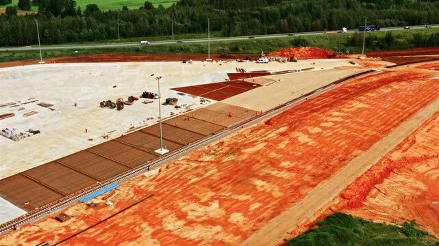 Aerial view of a large construction site featuring red soil, concrete foundations, and rebar grids with workers present near a highway in Latvia.