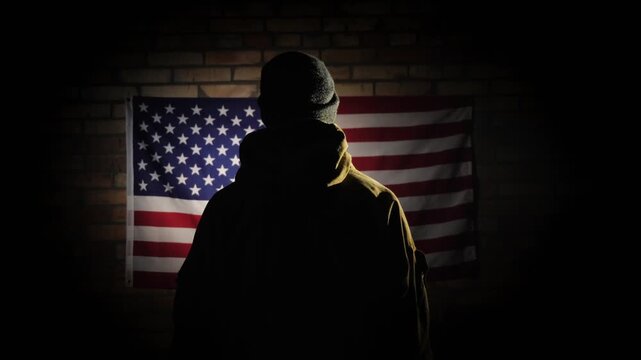 Silhouette of man standing before wall mounted american flag in dimly lit room.
