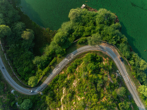 Aerial view of a winding road along the lush green banks of a lake with rocky terrain in Udaipur, Rajasthan, India.