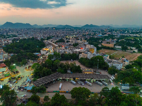 Aerial view of the bus terminal with parked buses surrounded by urban buildings, lush trees, and distant hills under a twilight sky in Udaipur, Rajasthan, India.