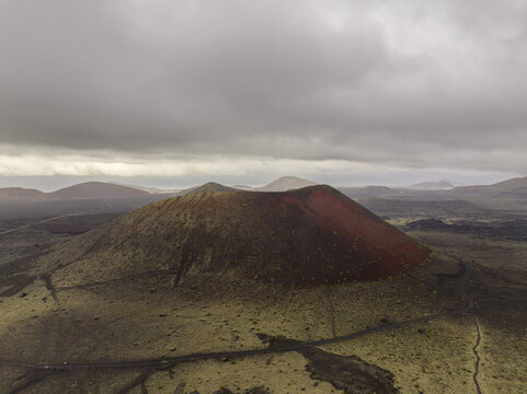 Aerial view of a striking red and brown volcanic crater surrounded by vast lava fields and rolling hills under a dramatic cloudy sky in Lanzarote, Canarias, Spain.