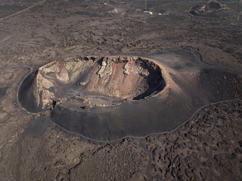 Aerial view of a volcanic crater with rugged dark lava fields, reddish rock formations, and a winding path around the rim in Lanzarote, Canarias, Spain.