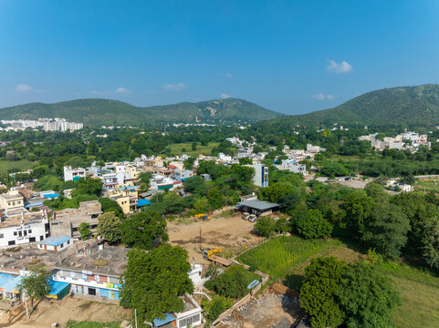 Aerial view of a residential area in Udaipur, Rajasthan, India, featuring scattered buildings, lush green trees, and rolling hills in the background under a clear blue sky.
