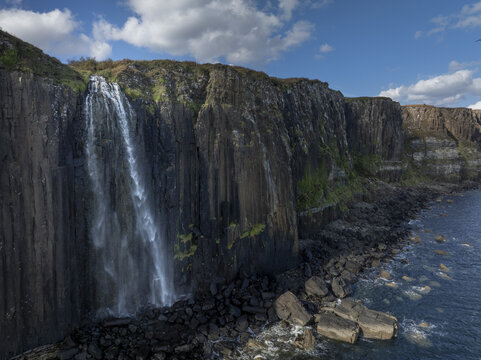 Aerial view of Mealt Falls cascading down dramatic basalt cliffs into the sea, with rocky shoreline and blue sky in Scotland, United Kingdom.