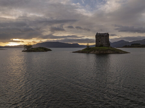 Aerial view of Castle Stalker situated on a small island in a loch surrounded by mountains under a dramatic sunset sky in Scotland, United Kingdom.