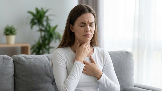 A young woman sitting on a grey sofa feeling unwell, touching her sore throat and chest, suffering from a cold, flu, or respiratory health problem