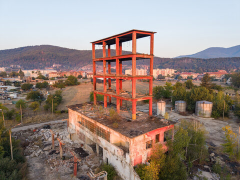 Aerial view of an abandoned industrial structure with a red steel frame rising above a derelict concrete building, surrounded by trees and mountains in Velingrad, Pazardzhik, Bulgaria.