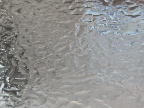Extreme close-up macro photography of clear water droplets and condensation forming on a smooth metallic or glass surface. Features a shallow depth of field with soft bokeh, highlighting the natural