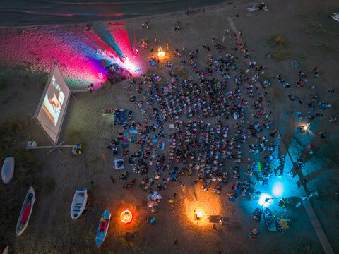 Aerial view of the Quarantine Film Festival on the beach at night with a large crowd watching a movie, colorful stage lights, and small boats in Asparuchovo, Varna, Bulgaria.