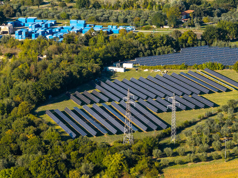 Aerial view of a solar farm with rows of photovoltaic panels and electricity pylons in a rural landscape with trees and blue storage containers in Nera Montoro, Umbria, Italy.