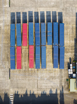 Aerial view of rows of blue and red shipping containers arranged in a concrete yard with long shadows cast by the afternoon sun in Taiwan.