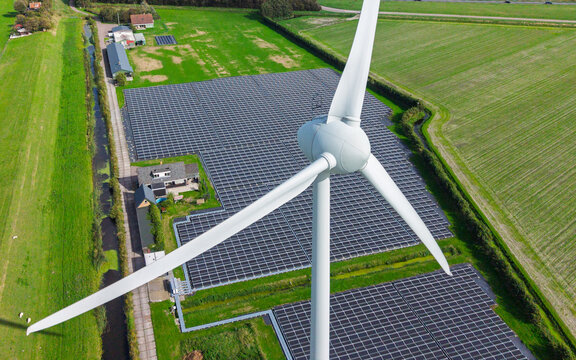 Aerial view of a large wind turbine standing next to a vast field of solar panels in a rural landscape with green fields and a small canal in 't Zand, North Holland, Netherlands.