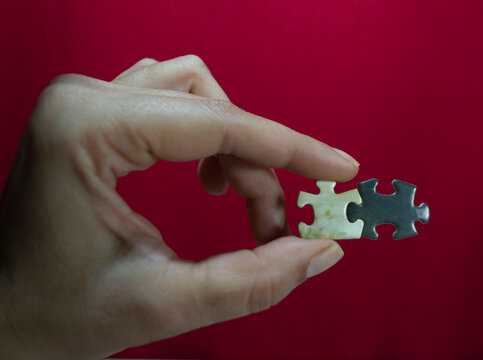 Close up of a hand holding two connected jigsaw puzzle pieces against a solid red background. Teamwork, connection, solution and partnership concept.