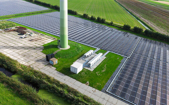 Aerial view of a wind turbine base surrounded by extensive solar panel arrays in a rural landscape with green fields and irrigation ditches in 't Zand, North Holland, Netherlands.