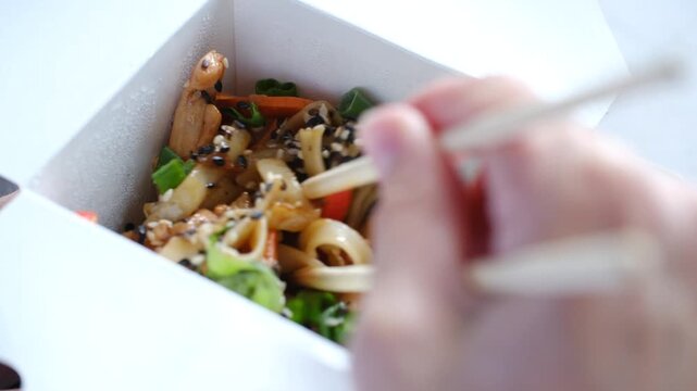 Close up person eating delicious asian udon noodles with chicken, vegetables, sesame seeds from white takeout box using chopsticks, representing convenient and tasty lunch option