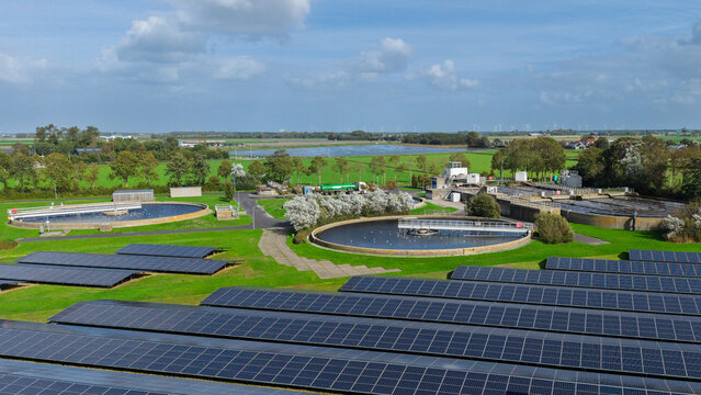 Aerial view of a water treatment plant with circular sedimentation tanks, solar panel arrays, and green fields in Schagerbrug, Noord-Holland, Netherlands.