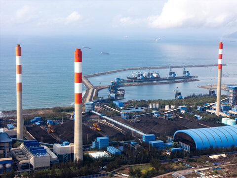 Aerial view of a coal-fired power plant with tall red and white chimneys, coal storage piles, industrial buildings, and a harbor along the coastline in Cilacap, Central Java, Indonesia.