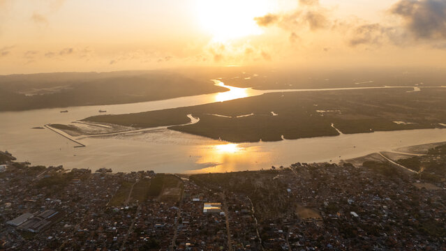 Aerial view of the river mouth and coastal landscape at sunset with golden light reflecting on the water, surrounded by urban rooftops and mangrove forests in Cilacap, Central Java, Indonesia.
