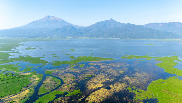 Aerial view of Lake Rawa Pening with lush green vegetation, water channels, and distant mountains under a clear blue sky in Waduk Rawapening, Central Java, Indonesia.