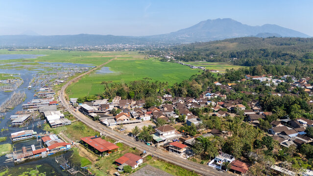 Aerial view of Lake Rawa Pening, featuring a village, green rice fields, and floating structures along the water, with mountains in Waduk Rawapening, Central Java, Indonesia.