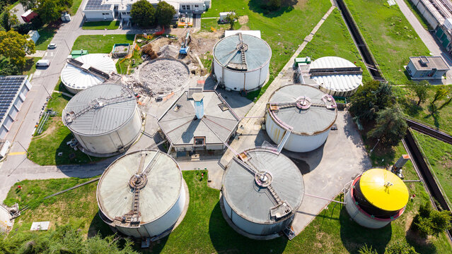 Aerial view of a water treatment plant featuring large circular sedimentation tanks, a central hexagonal building, and surrounding green lawns in an industrial site, Milan, Lombardy, Italy.