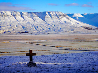A solitary stone cross headstone overlooks a vast, snow-dusted valley toward massive snow-covered basalt mountains under a clear blue sky in rural Iceland © Gregory