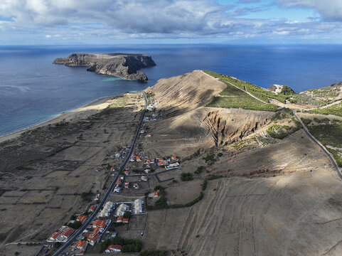 Aerial view of Ponta da Calheta, featuring rugged coastal cliffs, a sandy beach, scattered houses, and the Atlantic Ocean in Porto Santo, Madeira, Portugal.