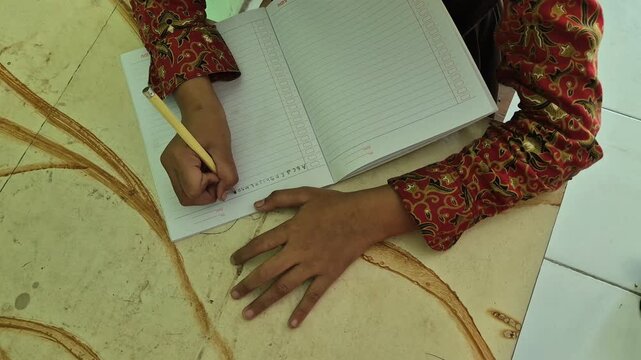 Close up of a person wearing a batik shirt writing in a notebook with a yellow pencil